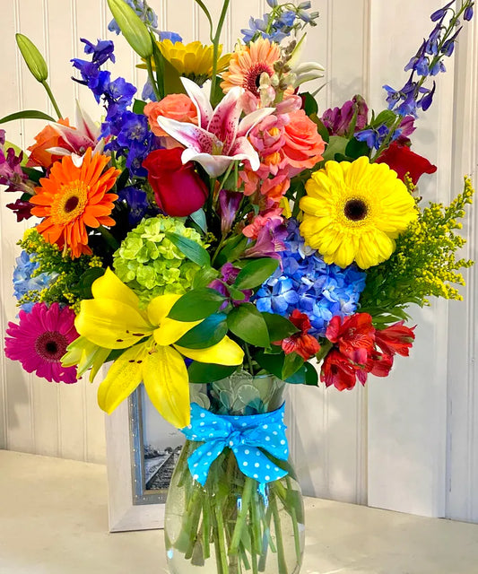 Vase of yellow lilies, green hydrangeas, gerbera daisies & blue delphinium on a round table inside a florist in clayton, nc
